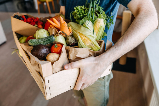 Közelkép az emberről, aki egy dobozt tart tele különböző organikus zöldségekkel, gyümölcsökkel és bogyókkal. Élelmiszerszállítási koncepció. - Fotó, kép