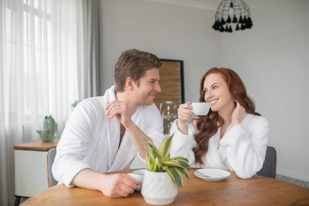 A couple in white bathrobes having coffee and feeling happy - Foto, imagen