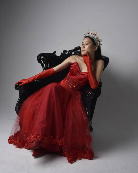 Ganztägiges Porträt einer schönen jungen Asiatin mit rotem Korsett, langen Opernhandschuhen und einer verzierten gotischen Königinnenkrone. Anmutige Pose im Sitzen auf Stuhl, isoliert auf Studiohintergrund. - Foto, Bild