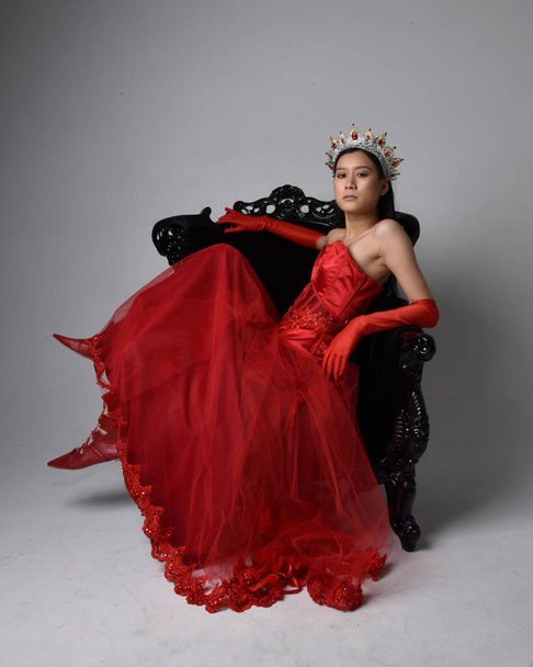 Retrato de comprimento total de bela jovem asiática vestindo espartilho vermelho, luvas de ópera longa e coroa rainha gótica ornamentada. Graciosa posando enquanto sentado na cadeira, isolado no fundo do estúdio. - Foto, Imagem