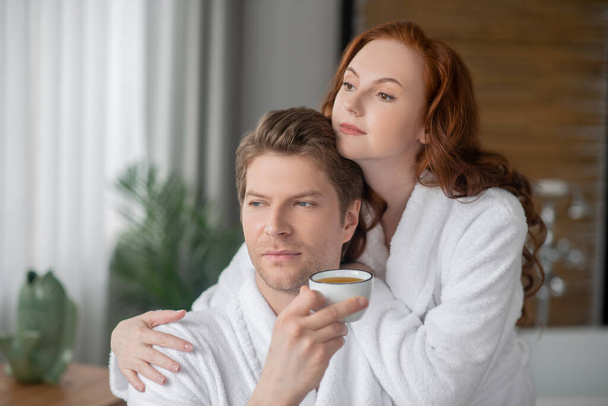 Ginger woman in a white bathrobe hugging her husband and looking dreamy - Foto, afbeelding