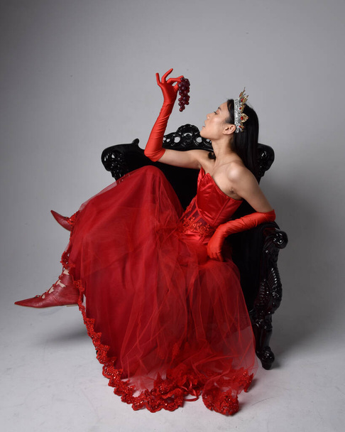 Full length  portrait of beautiful young asian woman wearing red corset, long opera gloves and ornate gothic queen crown. Graceful posing while seated on chair, isolated on studio background. - Foto, imagen