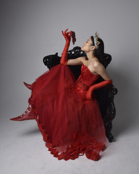 Full length  portrait of beautiful young asian woman wearing red corset, long opera gloves and ornate gothic queen crown. Graceful posing while seated on chair, isolated on studio background. - Fotografie, Obrázek