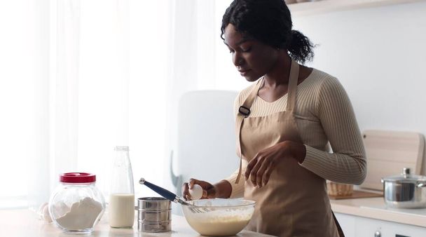 Jovem senhora afro-americana vestindo avental quebrando ovo e adicionando a outros ingredientes, preparando massa na cozinha - Foto, Imagem