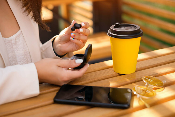 Žena vytáhne bezdrátová bluetooth sluchátka z nabíjecího pouzdra. Na stole je smartphone, sluneční brýle a žlutý šálek kávy. Čas poslouchat hudbu a odpočívat. sluchátka jsou moderní - Fotografie, Obrázek