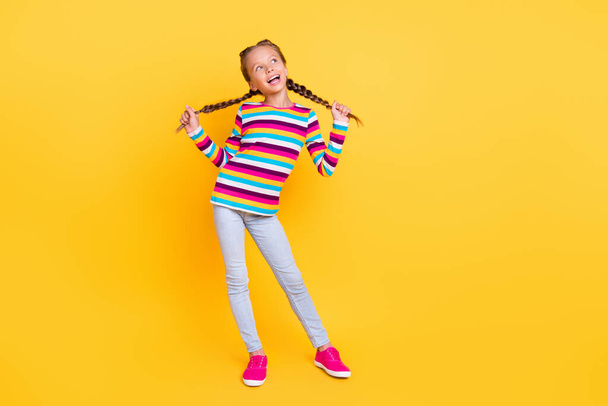 Photo of cheerful small lady play braids look empty space wear striped shirt jeans shoes isolated yellow color background - Photo, image