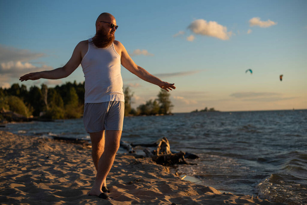 A humorous portrait of a brutal man in a T-shirt and boxers on the beach at sunset - Foto, Imagen
