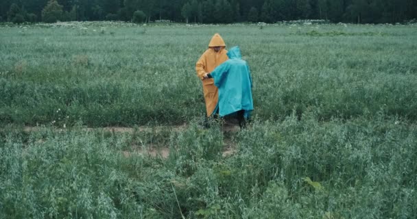 Man in gele regenjas en vrouw in blauwe regenjas houden elkaars hand vast en dansen in het veld - Video