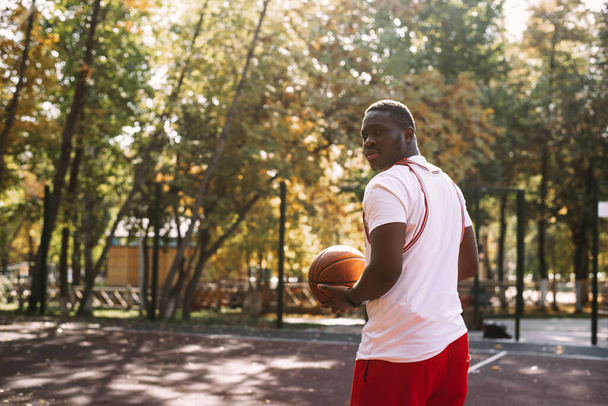 Genç bir Afrikalı Amerikalı adam açık havada basketbol oynuyor, gülümsüyor ve kameraya bakıyor. Açık hava sporları - Fotoğraf, Görsel