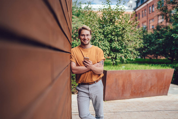 Smiling male in glasses and orange shirt and gray pants looking at camera while standing with crossed arms on street leaning on wooden brown facade of modern building and interacting with smartphone against blurred green trees - Фото, зображення