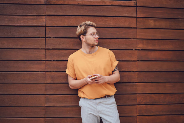 Σοβαρός μοντέρνος τύπος με πορτοκαλί πουκάμισο και ανοιχτό γκρι παντελόνι κρατώντας το κινητό τηλέφωνο στα χέρια και κοιτάζοντας μακριά, ενώ στέκεται δίπλα σε κόκκινη σανίδα ξύλινο τοίχο - Φωτογραφία, εικόνα