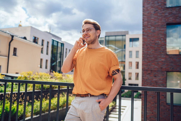 From below of successful modern guy in orange shirt having phone conversation and smiling happy with good news while standing on pedestrian bridge in city  - Фото, зображення