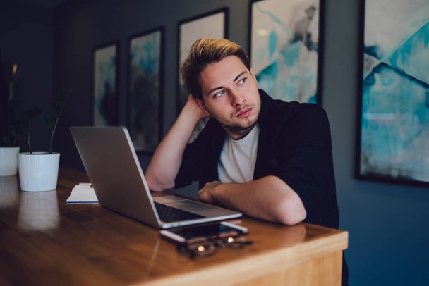 Junger Mann im lässigen Outfit kratzt am Kopf und schaut weg, während er am Tisch sitzt und während der Arbeit im modernen Büro nachdenkt - Foto, Bild