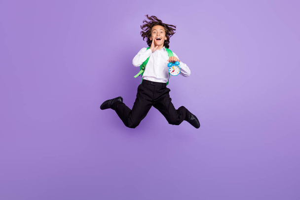 Full size photo of impressed small brunet boy jump hold clock wear bag shirt trousers sneakers isolated on purple background - 写真・画像