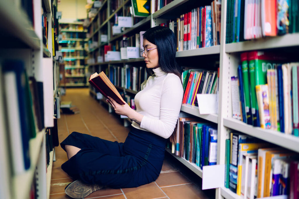 Side view of young pretty focused female in glasses sitting cross-legged on floor between bookcases in library and reading book - Fotografie, Obrázek
