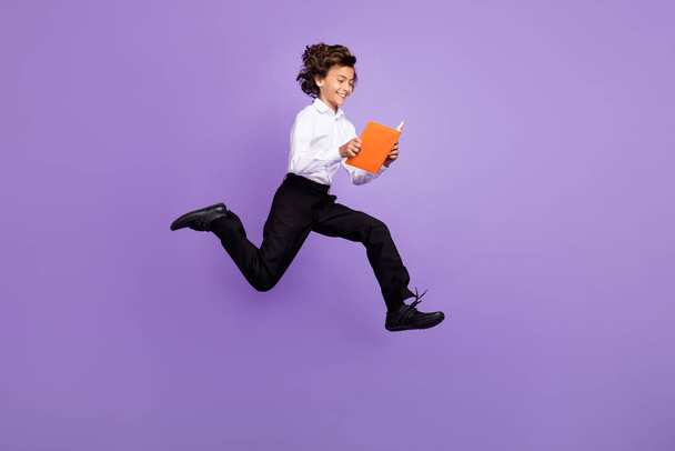 Full size photo of nice little brunet boy jump read book wear shirt trousers sneakers isolated on purple color background - Fotografie, Obrázek