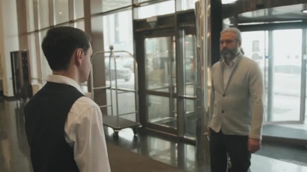 若い男性受付で歓迎されている現代のホテルにスーツケースを歩くと成熟したビジネスマンのスローモー追跡ショットで制服 - 映像、動画