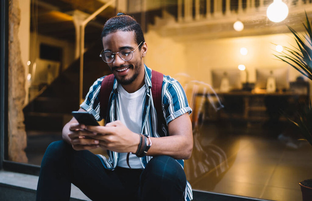 Smiling african american hipster guy checking via smartphone application sitting on street during free time,positive dark skinned male satisfied with 4G connection for browsing and share media content - Foto, Bild