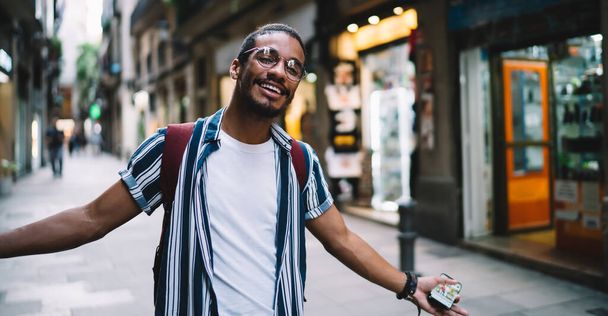 Sırt çantalı, gözlüklü, akıllı telefonlu neşeli siyah saçlı adam yaz günü caddede yürürken kollarını akıllı telefonuyla açıyor. - Fotoğraf, Görsel