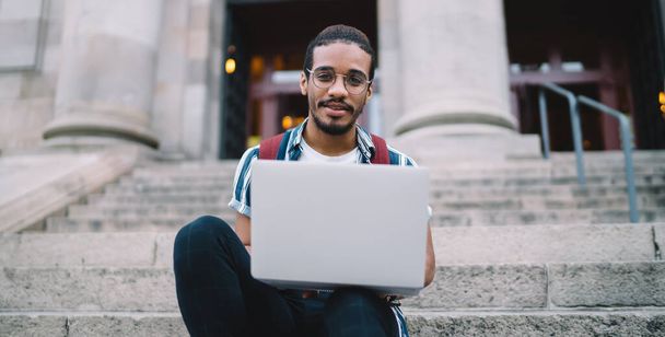 Half length portrait of handsome prosperous african american man looking at camera sitting near university using technology for learning, smiling dark skinned male blogger use laptop on freelance job - Fotoğraf, Görsel