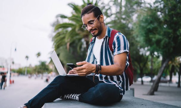 Young millennial dark skinned guy holding mobile phone reading information about online booking, hipster guys spending time time outdoors holding mobile phone working via laptop on freelance - Photo, Image