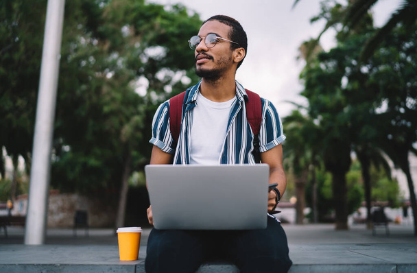 Cool ethnic guy in trendy summer wear and eyeglasses browsing netbook and drinking coffee to go while working remotely on street and looking away attentively - 写真・画像