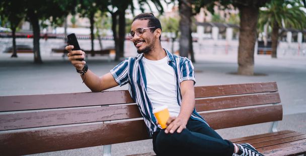 Cheerful african american hipster guy communicating in social networks sitting on street satisfied with 4G connection, positive dark skinned male enjoying video call on smartphone on free time - Photo, image