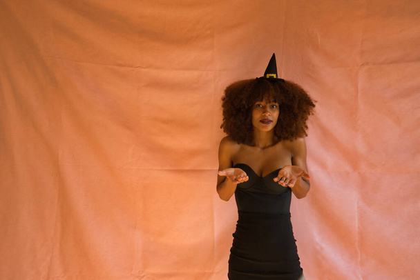 молодая латиноамериканка, одетая как ведьма с ладонями вверх, как будто колеблется на розовом фоне на Хэллоуин. - Фото, изображение