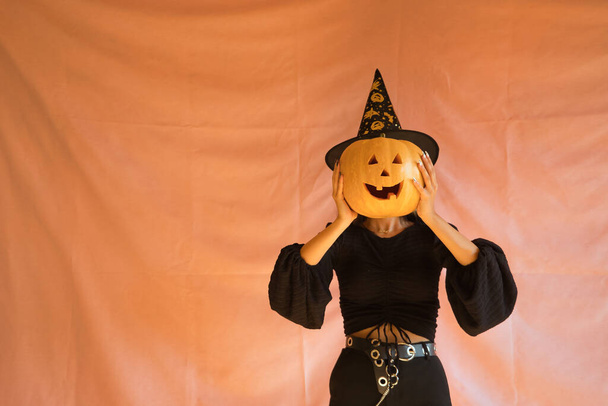 Mladá hispánka oblečená jako čarodějnice držící dýni před obličejem na růžovém pozadí pro Halloween. - Fotografie, Obrázek