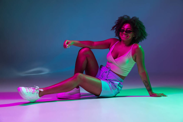 Portrait of African young girl posing isolated on dark blue studio background in pink neon light. Concept of human emotions, facial expression, youth, sales, ad. - 写真・画像