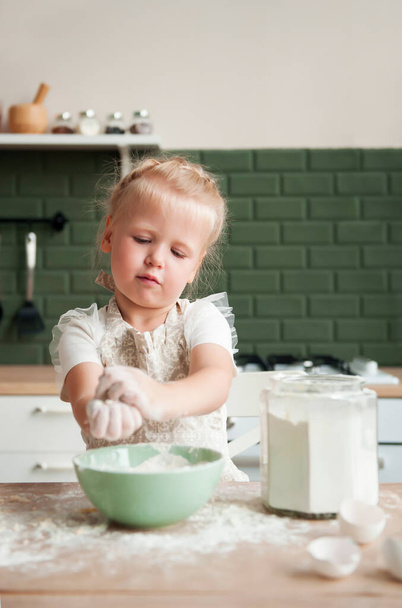 Το μικρό χαρούμενο κορίτσι ετοιμάζει λεφτά στην κουζίνα. Το όμορφο παιδί μαγειρεύει στην κουζίνα. - Φωτογραφία, εικόνα