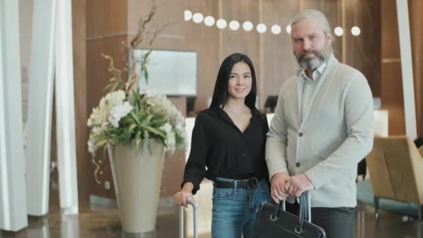 Retrato lento médio de casal moderno posando para câmera com malas em pé em hotel elegante com recepção em fundo embaçado - Filmagem, Vídeo