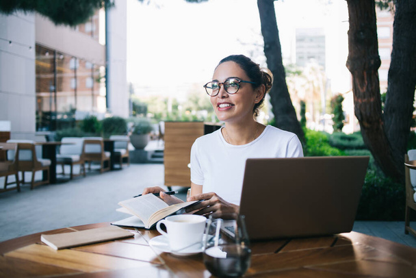 Young cheerful female in casual clothes and eyeglasses sitting at table with laptop and copybook while working in soft daylight - Foto, Imagem