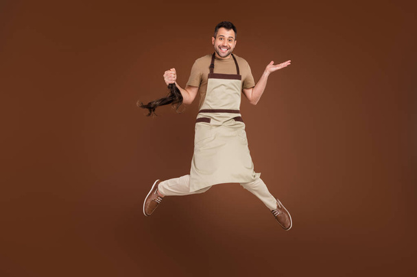 Photo of sweet funny young guy dressed beige uniform jumping cutting ponytail isolated brown color background - Фото, изображение