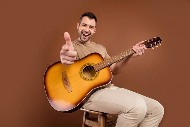 Photo portrait brunet man in pastel t-shirt playing guitar pointing finger at you isolated brown color background - Fotografie, Obrázek