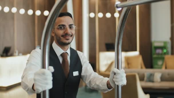 Medium slowmo portret van lachende jonge mannelijke piccolo in uniform poseren voor camera met bagage kar staan in chique hotel lobby - Video