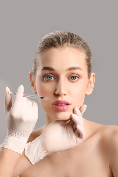 молодая женщина получает инъекцию наполнителя в губы на сером фоне - Фото, изображение