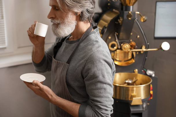 Senior man met kopje warme drank in de buurt van moderne koffiebranderij - Foto, afbeelding