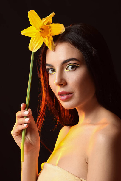 Hermosa mujer joven con maquillaje elegante y flor sobre fondo oscuro - Foto, imagen