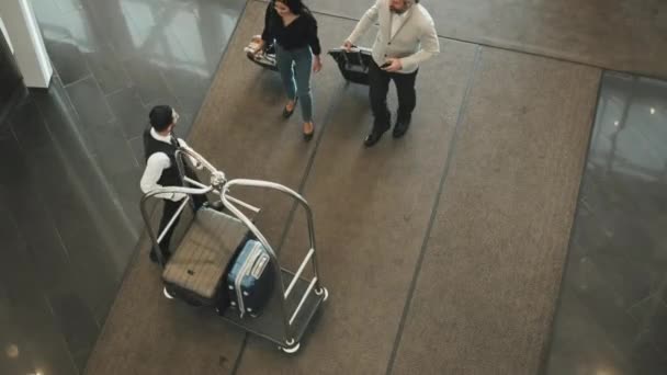 Vista superior de la pareja turística moderna que llega al hotel de clase alta dando su equipaje a un joven portero masculino que lo coloca en el carrito - Metraje, vídeo