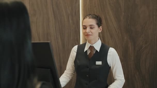 Μεσαία βολή της νεαρής γυναίκας ρεσεψιονίστ μιλώντας ευγενικά με την φιλοξενούμενη γυναίκα κατά το check-in, όπως ψάχνει για τα διαθέσιμα δωμάτια στο ξενοδοχείο - Πλάνα, βίντεο