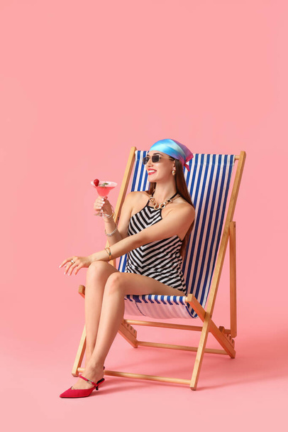 Beautiful young woman with cocktail sitting on deck chair against color background - Fotografie, Obrázek