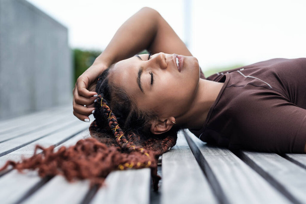 Portrait of young woman lying on a wood bench. Braid, mixed race. She is wearing sportswear. - Fotoğraf, Görsel