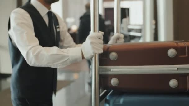 Midsection tiro de porteiro masculino irreconhecível em uniforme e luvas transportando carrinho de bagagem através do lobby do hotel moderno - Filmagem, Vídeo