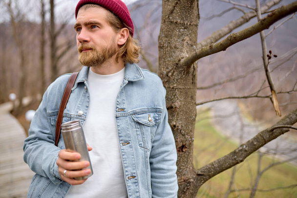 Thoughtful Caucasian Male Traveler Standing With Thermos Next To Tree In Forest - Foto, Imagen