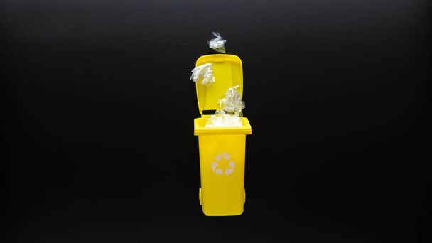 Trash plastic bin. Yellow dustbin for recycle plastic trash on black background. Container for disposal garbage waste and save environment. Flat lay - Photo, Image