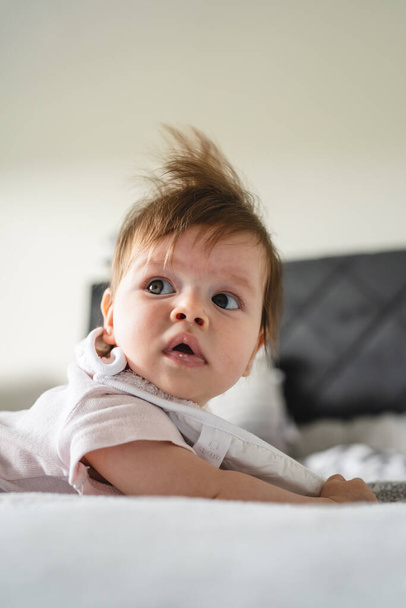 Маленький белый ребенок четыре месяца лежит на кровати дома в светлой комнате в день с копировальным пространством глядя на боковой живот вниз - Фото, изображение