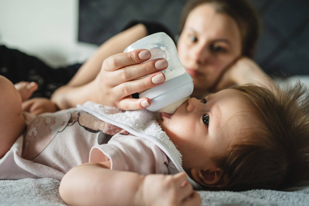 Közelkép kis kaukázusi csecsemő négy hónapos feküdt az ágyon otthon, miközben az anyja táplálja őt egy üveg formula tej anyaság és a szülői koncepció valódi emberek - Fotó, kép