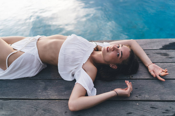 From above of alluring sensual female in white wear relaxing on poolside and looking at camera while spending time in resort - 写真・画像
