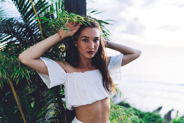 Young slender woman in white crop top touching foliage and confidently looking at camera while lounging in park near sea - Foto, Imagem
